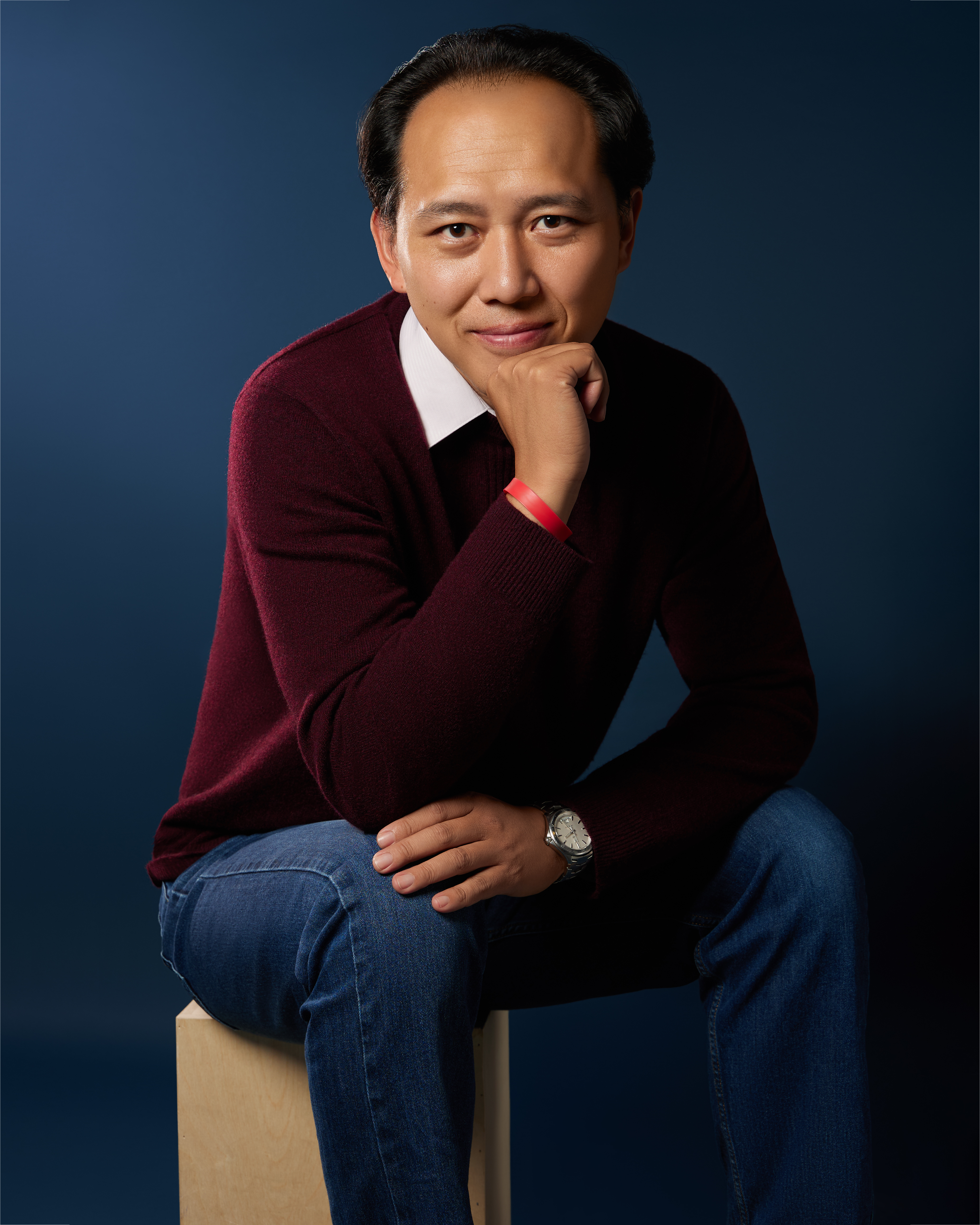 Haowen is a Taiwanese man with wavy black hair, brown eyes, and a light-medium complexion. Sitting with one arm resting on his knees and the other on his chin, he is smiling intently at the camera. He is in a dark blue backdrop and wearing a wine-red sweater, blue jeans, with a wristband and a silver wristwatch.