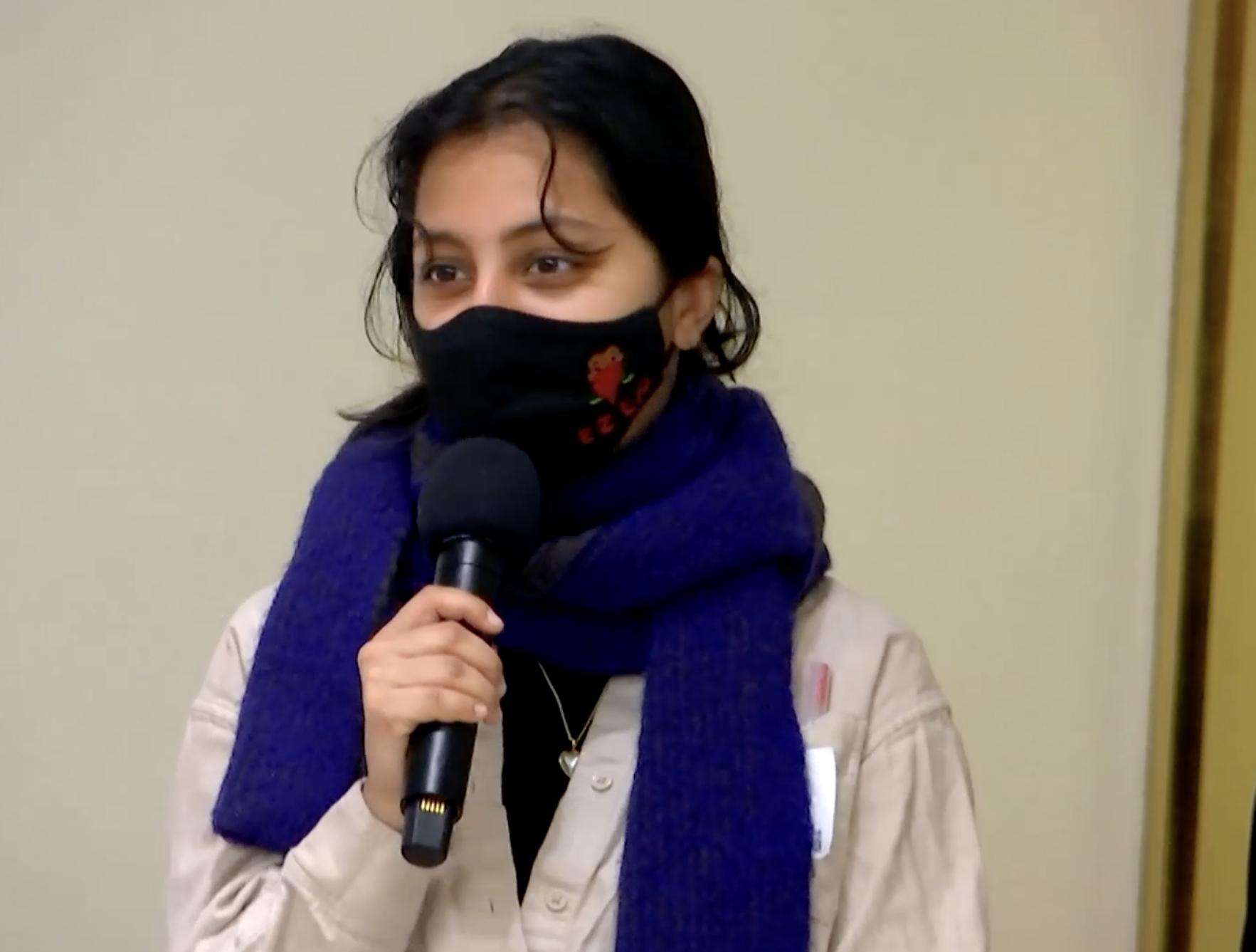 South asian person with hair pulled into a bun, wearing a black mask with a zapatista image and a thick blue scarf wrapped around their neck, holding a mic to their face