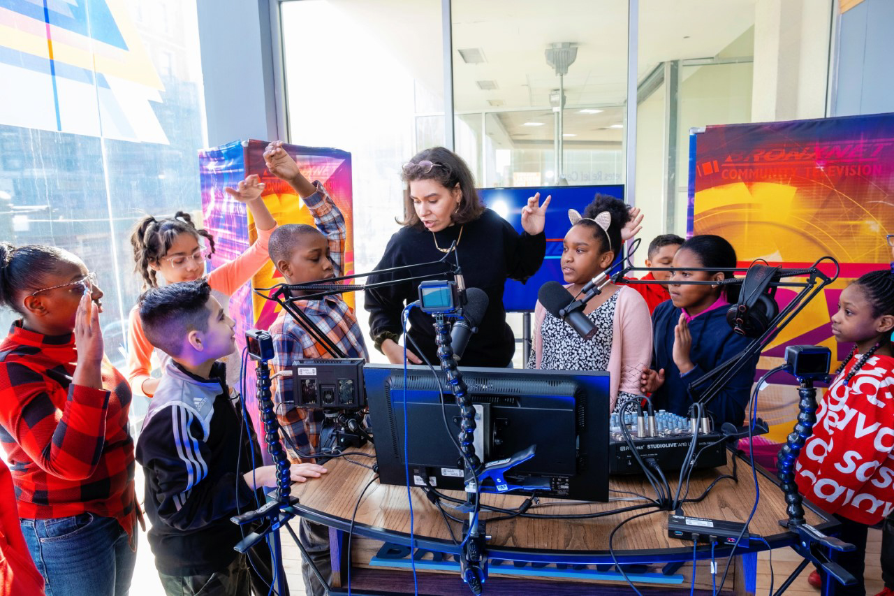 A group of children and one adult gather behind a soundboard setup that includes a monitor and multiple microphones. The children look at the adult as she raises her hand in the air; a few children mimic the pose.