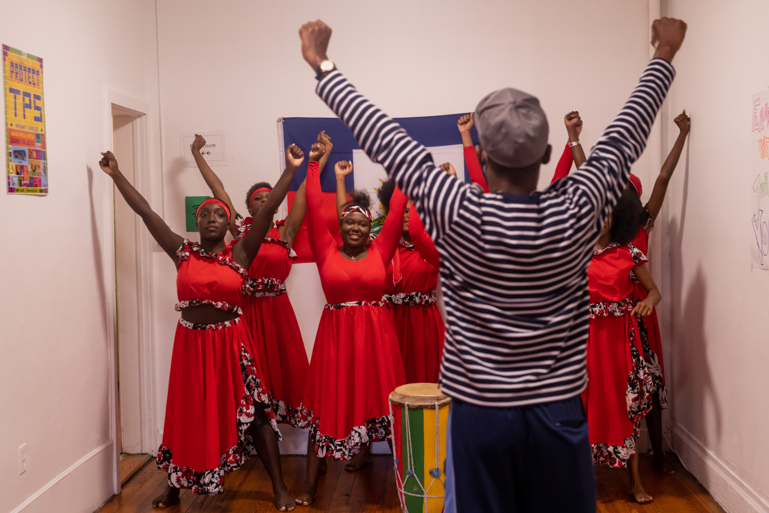 A group of people stand in a white room wearing red shirts and skirts trimmed with a white, red, and black floral print. Each person stands with their arms over their heads in a