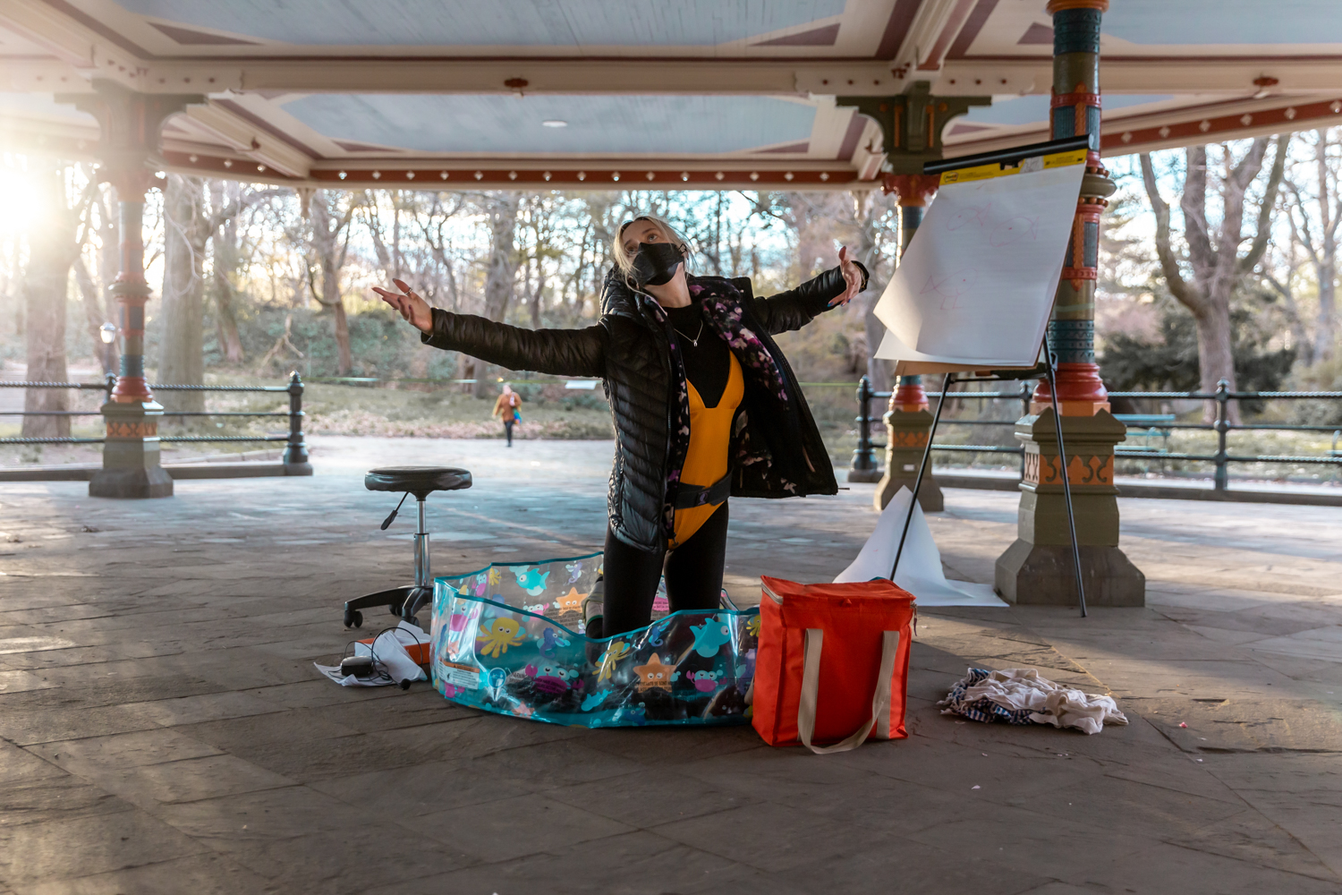 An adult, wearing an orange leotard over a black unitard and black puffy coat, kneels in an empty baby pool. Their upturned face is partially covered with a black surgical mask and their arms are outstretch on either side of their body. They are surrounded by various objects such as a stool and easel.