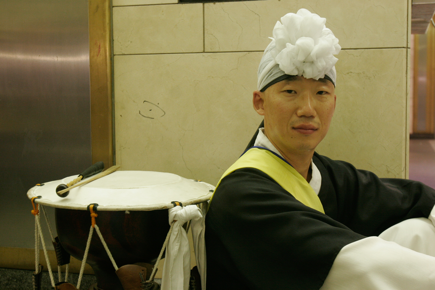 A close up of a musician and their drum. The musician sits on the right with their back resting on the drum to the left. They sit in profile, but turn their head is turned towards the camera. They wear a headdress made of a delicate white fabric arranged to look like a flower on the top of their forehead.