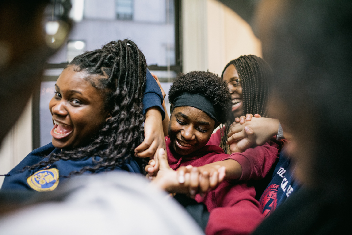 A close up of three smiling people framed between the blurred faces of a crowd. They contort their bodies in order to interlock arms. 