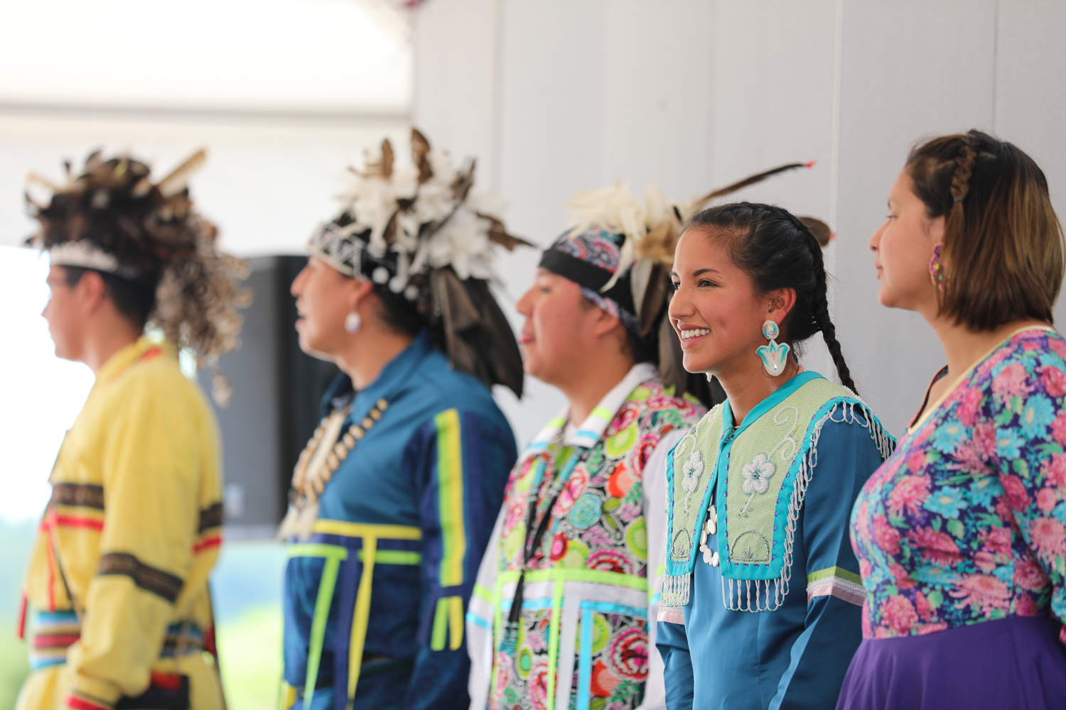 A group of indigenious folks stand diagonally in a row, looking away from the camera. Some appear to be talking or singing, but each face gets slightly blurrier as they move away from the camera. The participants are formally dressed, some wearing headdresses or elaborate beadwork.