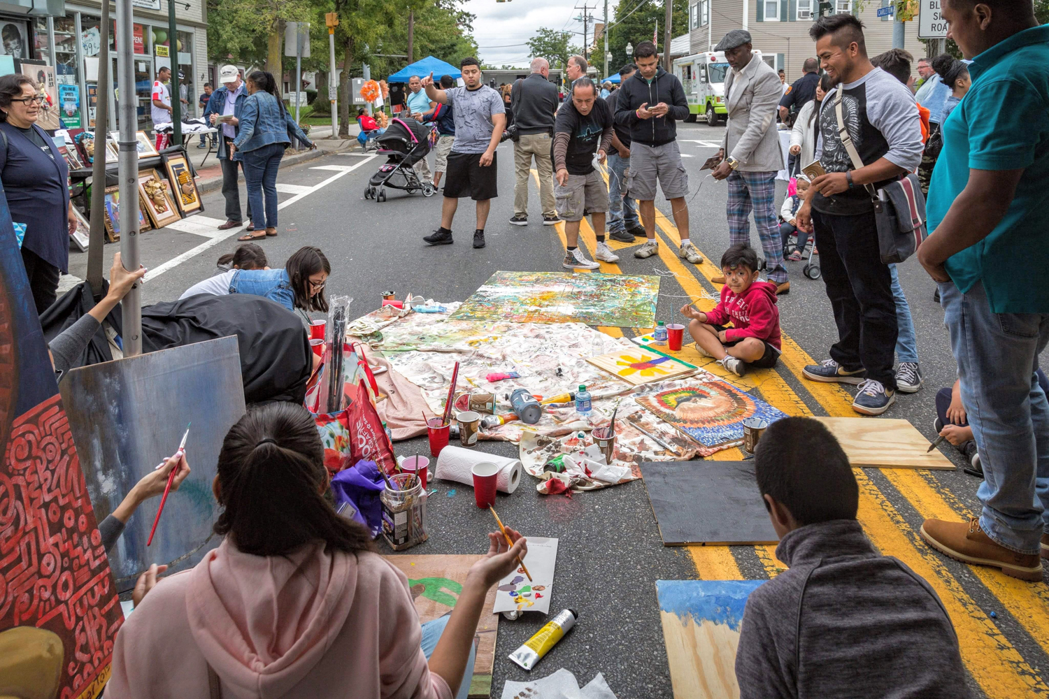 A street fair where a group of children have gathered to paint in the middle of the road. They are seated on the ground with paint supplies scattered every which way. A group of adults gather around smiling and admiring the artwork. 