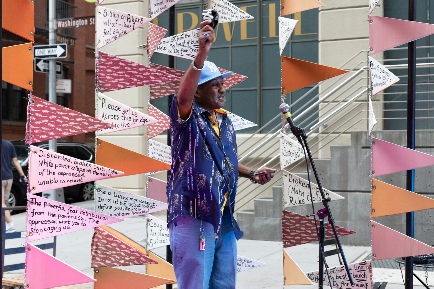 A Black man stands with their hand raised in the air. Behind him are strands of triangular shaped flags strung together in a vertical orientation. Some of the flags have handwritten text.